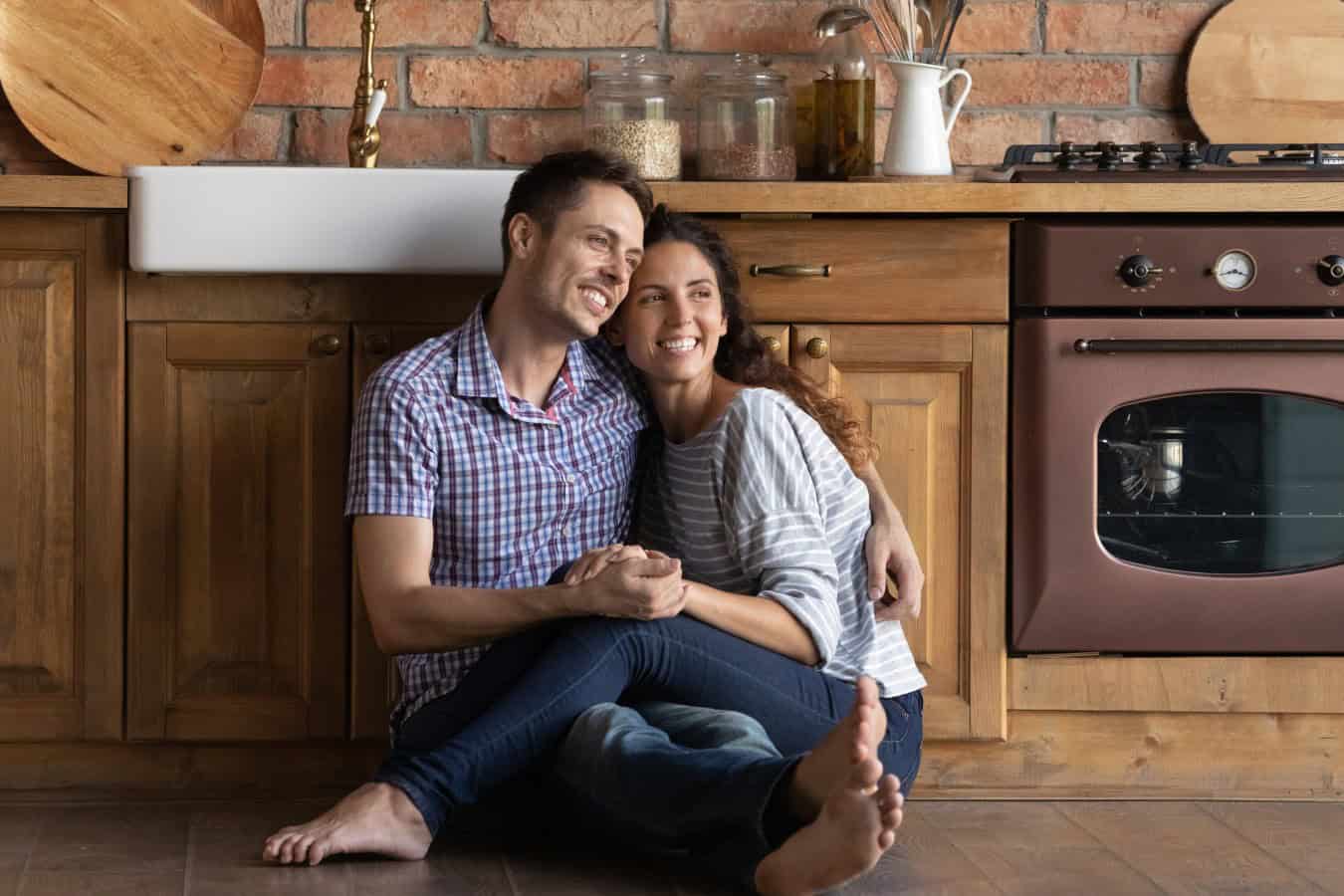 Overjoyed millennial couple renters sit on warm kitchen floor hugging look in distance dreaming happy future together. Smiling young man and woman embrace plan imagine new beginning or perspectives.
