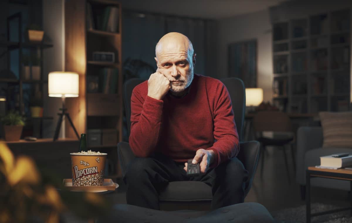 Bored senior man sitting on the armchair and watching TV, he is holding the remote control and changing channels