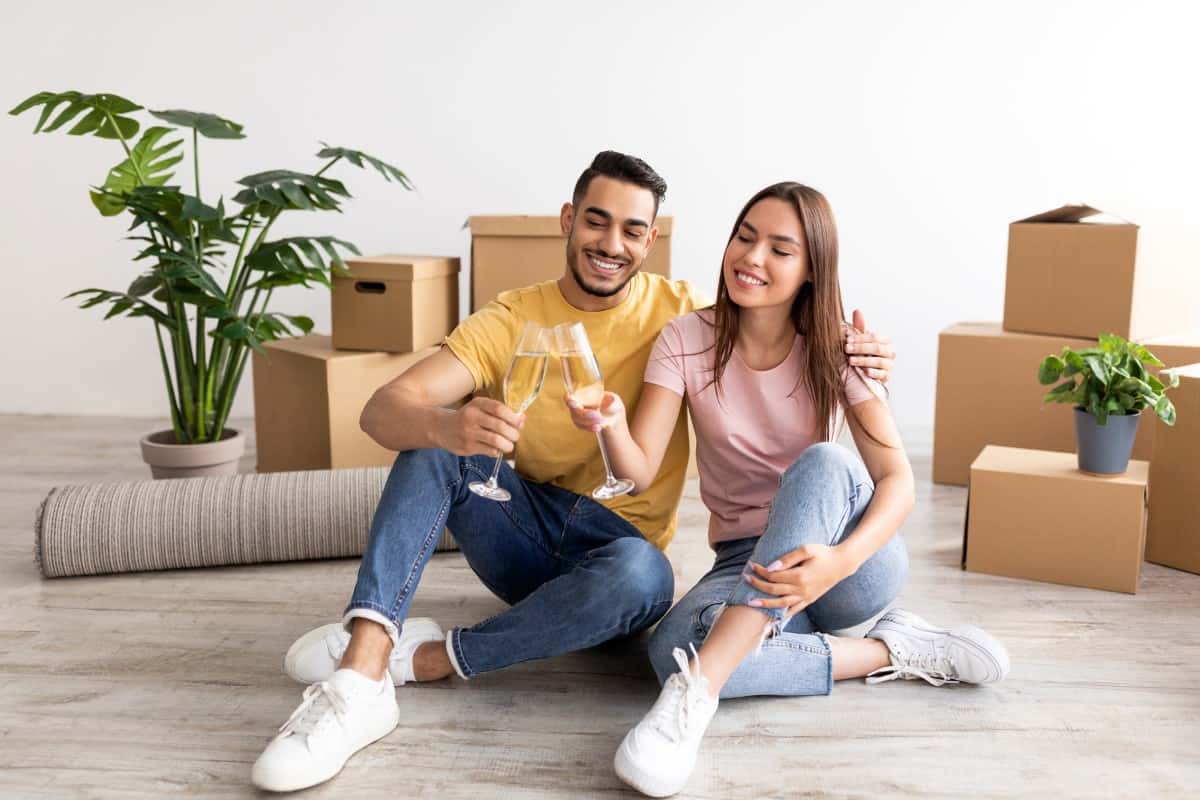 Millennial multiracial couple having housewarming party, sitting on floor with glasses of champagne, celebrating new home purchase, making toast among cardboard boxes. Moving day, relocation concept