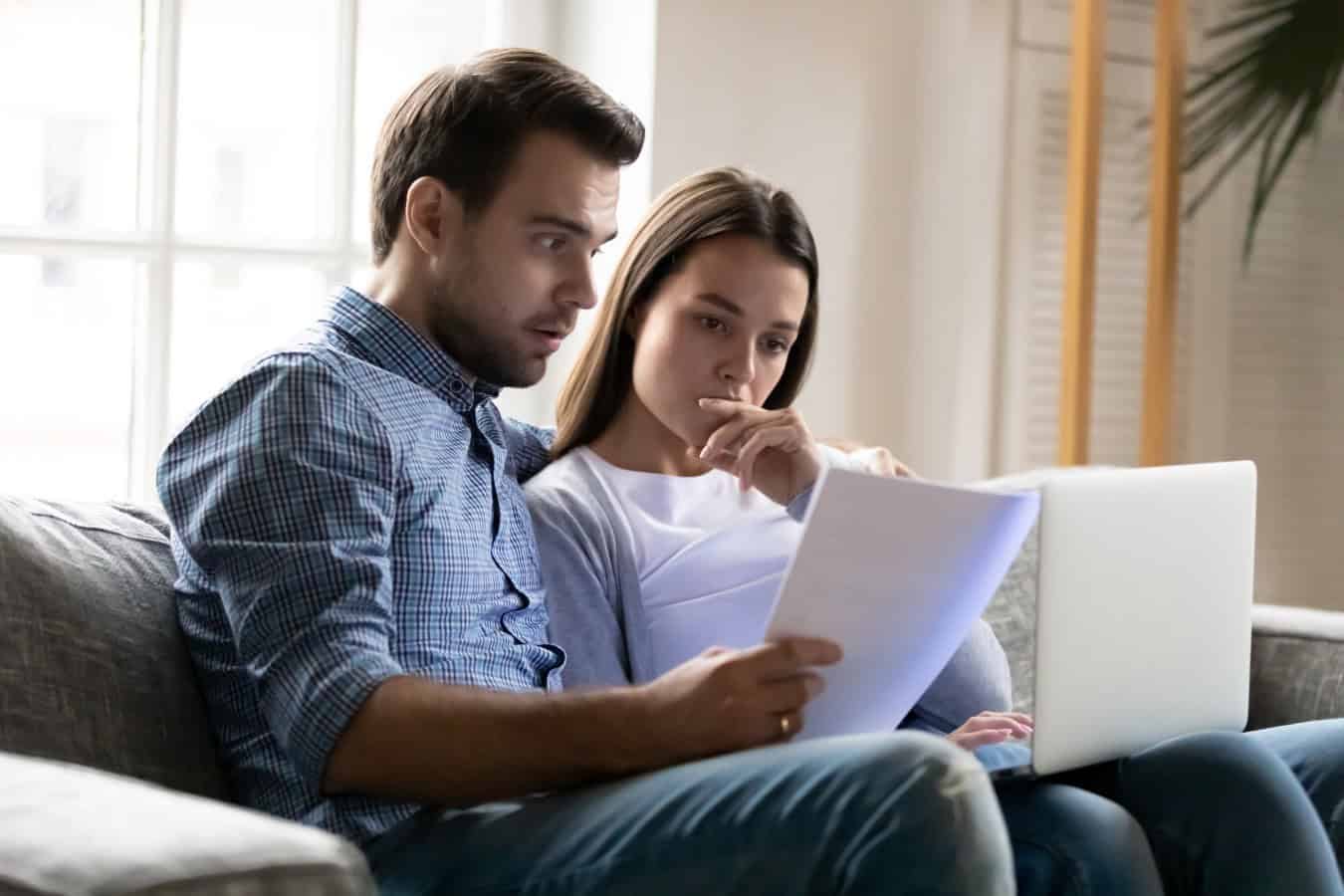 Shocked worried young couple reading documents, financial problems, using laptop, online banking service, checking mortgage or insurance contract terms, calculating domestic bills or taxes