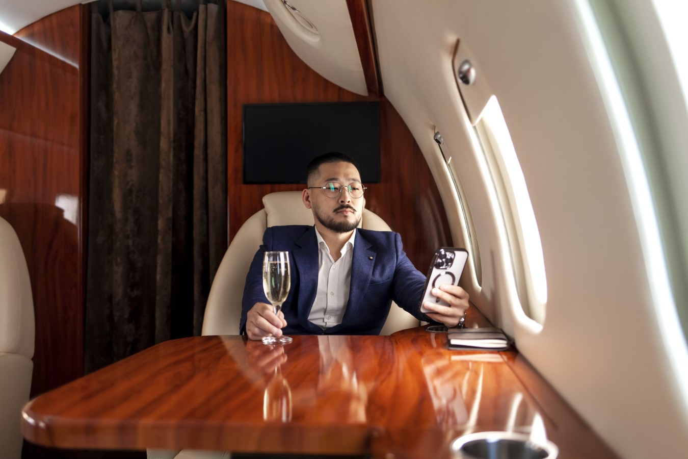 asian businessman in suit and glasses with glass of champagne flies in private luxury jet and uses smartphone, korean entrepreneur resting and looking at the phone in flight, luxury lifestyle