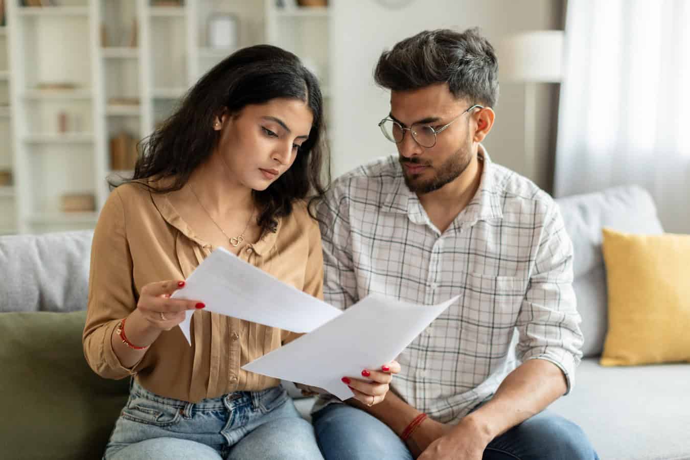 Indian couple checking bills, reading documents, unhappy man and woman holding papers, counting monthly spendings, sitting on sofa at home